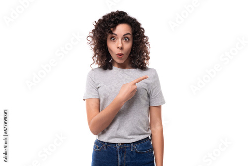 surprised european woman with black curly hair in a gray basic t-shirt points her fingers towards advertising space