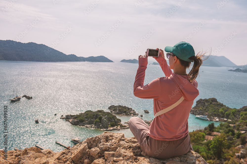 Beautiful traveler woman sitting on mountain peak looking at the sea water and making photo in summer