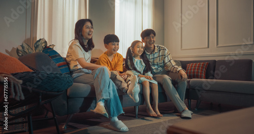 Fotografie Happy Korean Family At Home: Mother And Father Watching Funny Family Television Show And Laughing With Their Little Son And Daughter On The Couch