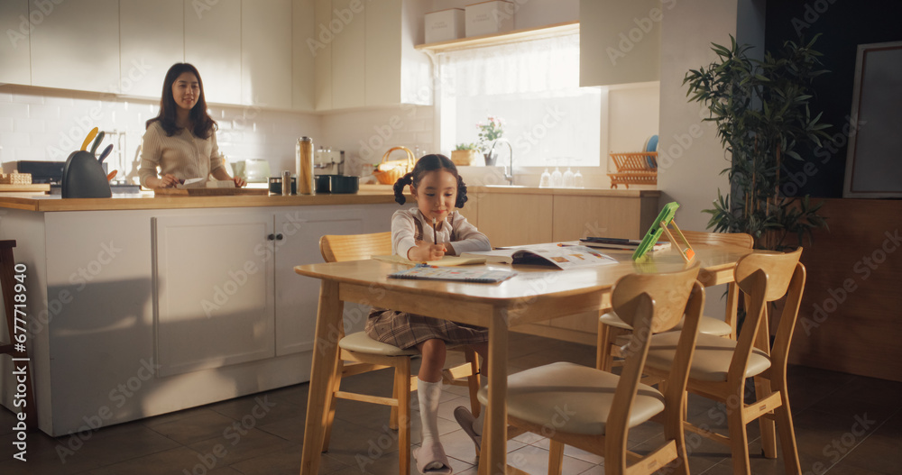 Portrait of a Korean Female Child Sitting at a Kitchen Table and Drawing While her Mother is Preparing Breakfast. Little Cute Girl Waiting for Nutritious Meal Before Going to School Portrait of a Korean Female Child Sitting at a Kitchen Table and Drawing While her Mother is Preparing Breakfast. Little Cute Girl Waiting for Nutritious Meal Before Going to School