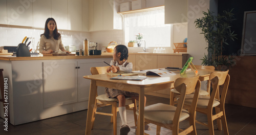 Portrait of a Korean Female Child Sitting at a Kitchen Table and Drawing While her Mother is Preparing Breakfast. Little Cute Girl Waiting for Nutritious Meal Before Going to School