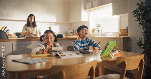 Portrait of a Korean Children Sitting at a Kitchen Table, Drawing and Writing While their Mother is Preparing Breakfast. Little Cute Kids Waiting for Nutritious Meal Before Going to School