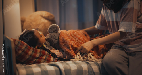 Korean Young Mother Caring for Her Lovely Daughter, Putting Her to Sleep in Bed at Home in the Evening. Babysitter Tucking Little Girl in Blanket, Making Sure She is Warm and Comfortable at Night photo