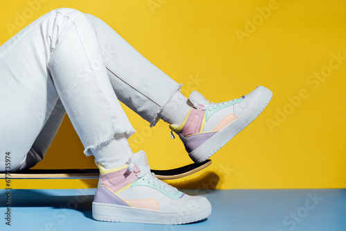Close up female legs in white jeans and retro style high-top multicolor sport sneakers shoes sitting on the skateboard on blue and yellow background. Vintage retro fashion style of 80s - 90s vibes. photo