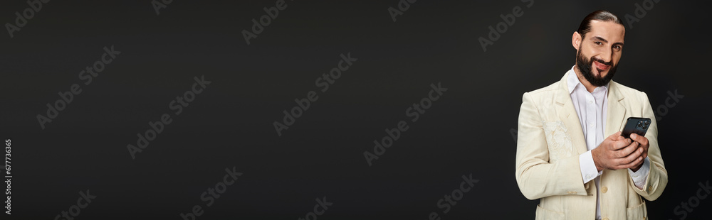 cheerful and bearded arabic man in white shirt and blazer texting smartphone on black, banner cheerful and bearded arabic man in white shirt and blazer texting smartphone on black, banner