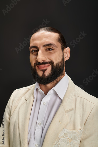 happy and bearded arabic man in white shirt and blazer looking at camera on black background