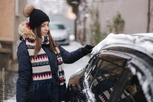 A woman is cleaning snowy window on a car with snow scraper. Pretty woman warmy dressed clean her car outdoors. Cold snowy and frosty morning. Black car photo