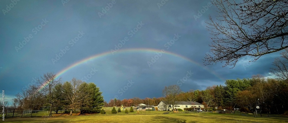Panoramic view of beautiful rainbow in blue sky over a house with lush green garden Panoramic view of beautiful rainbow in blue sky over a house with lush green garden