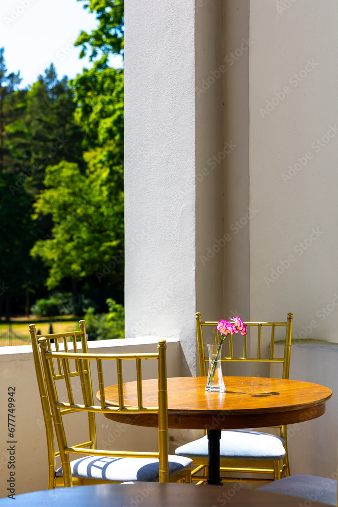 Empty tables on the terrace of a restaurant in the middle of a sunny day. In the blurred background, a view of the greenery of a city park. A vase with flowers on the table.