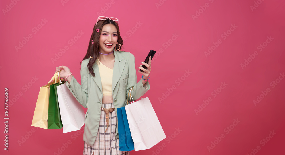 Trendy beautiful young Asian teen woman carrying colorful bags shopping online with mobile phone isolated on pink banner background with copy space. Trendy beautiful young Asian teen woman carrying colorful bags shopping online with mobile phone isolated on pink banner background with copy space.