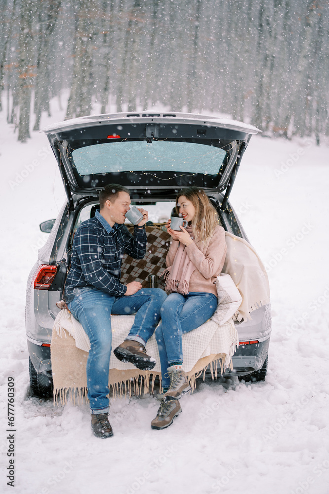 Couple drinking coffee from cups while sitting in the trunk of a car in a snowy forest