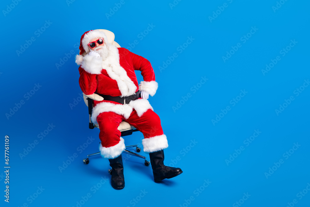 Full body photo of retired old man sit listen interested armchair wear trendy santa claus costume coat isolated on blue color background Full body photo of retired old man sit listen interested armchair wear trendy santa claus costume coat isolated on blue color background
