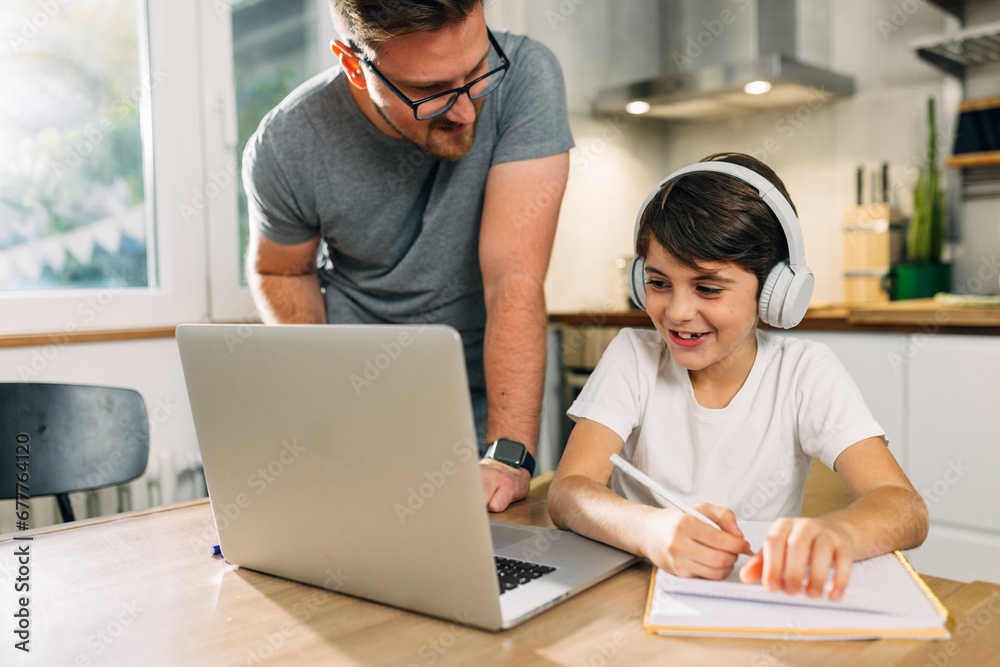 Happy kid is taking notes during the online class, nd his father helps him with answers. Happy kid is taking notes during the online class, nd his father helps him with answers.