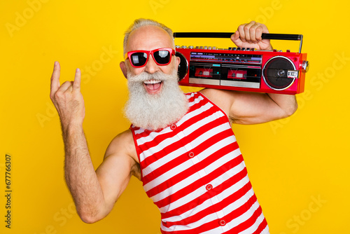 Portrait of cool funny pensioner feel young holding retro boombox showing rock n roll symbol fingers isolated on yellow color background