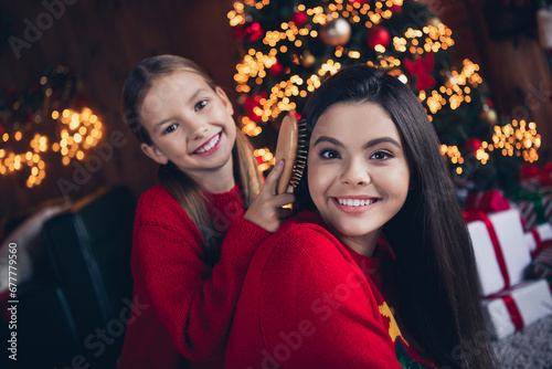 Photo of positive cute small sisters wear red jumpers doing xmas hairstyle brushing hair indoors new year north pole house