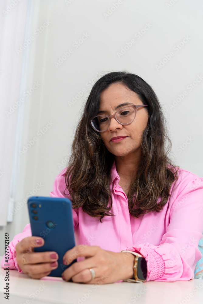 Portrait of a psychopedagogue, wearing pink clothes, in her office, sitting, looking at the appointments scheduled on her cell phone. Helps with school performance. Portrait of a psychopedagogue, wearing pink clothes, in her office, sitting, looking at the appointments scheduled on her cell phone. Helps with school performance.