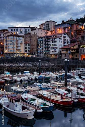 Vertical shot of the old town of Mutriku illuminated at dusk and the fishing port full of boats in the foreground, Gipuzkoa, Basque Country, Spain photo