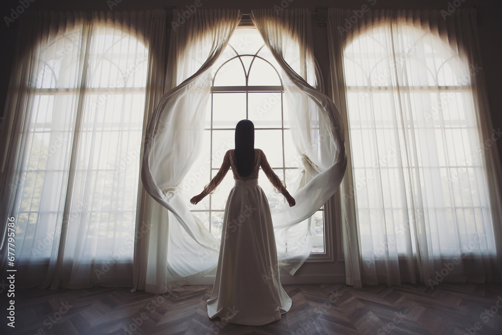 Bride near the window. the bride in a wedding dress opens the curtains of the window, stands back, you can see her silhouette, against the light, boudoir morning of the bride. wedding day. - obrazy, fototapety, plakaty 