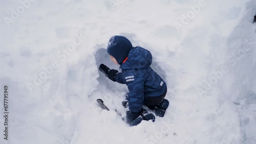 boy child in a warm jumpsuit drips snow in a snowdrift in winter with an iron shovel at home.