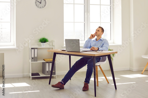 Young creative business man or corporate employee working in office, sitting at his desk with laptop computer and papers, writing business plan, holding hand with pen on chin and thinking © Studio Romantic