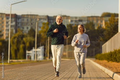 Fit senior couple enjoying jogging workout on sunny summer morning. Old male and female athletes running on pavement, with green trees and apartment buildings in background. Sport, exercise concept