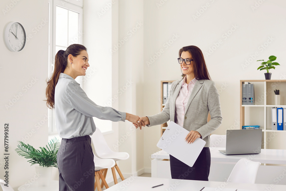 Young happy smiling business women standing in office on workplace shaking hands with each other reaching agreement and making a good deal or signing a contract. Business people at work. Young happy smiling business women standing in office on workplace shaking hands with each other reaching agreement and making a good deal or signing a contract. Business people at work.