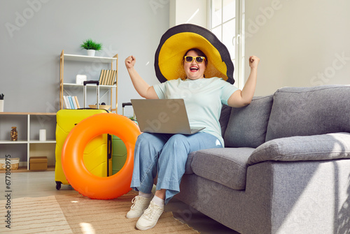 Funny fat plus size woman in summer sunhat and sunglasses sitting on sofa with laptop, suitcases and beach ring happy that she booked discount ticket or hotel room for vacation. Holiday travel concept photo