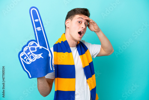 Young sports fan man isolated on blue background doing surprise gesture while looking to the side