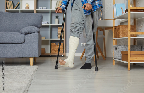 Man with fractured leg in plaster cast standing with crutches, selective focus. Cropped shot of plastered leg walking at home. Bone fracture, trauma, recovery, rehabilitation concept