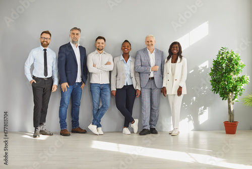 Group of well dressed business people posing together at wall in office. Smiling successful multiracial business team, corporate office colleagues standing close to each other looking at camera