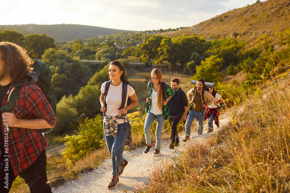 Active wanderlust people going hiking or trekking on good sunny day. Group of male female friends with backpacks following mountain trail route, leaving countryside with green woods and valleys behind - obrazy, fototapety, plakaty 