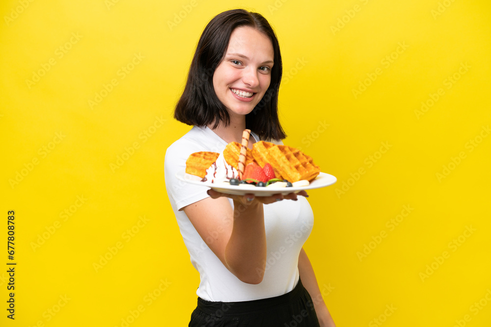 Young caucasian woman holding waffles isolated on yellow background with happy expression