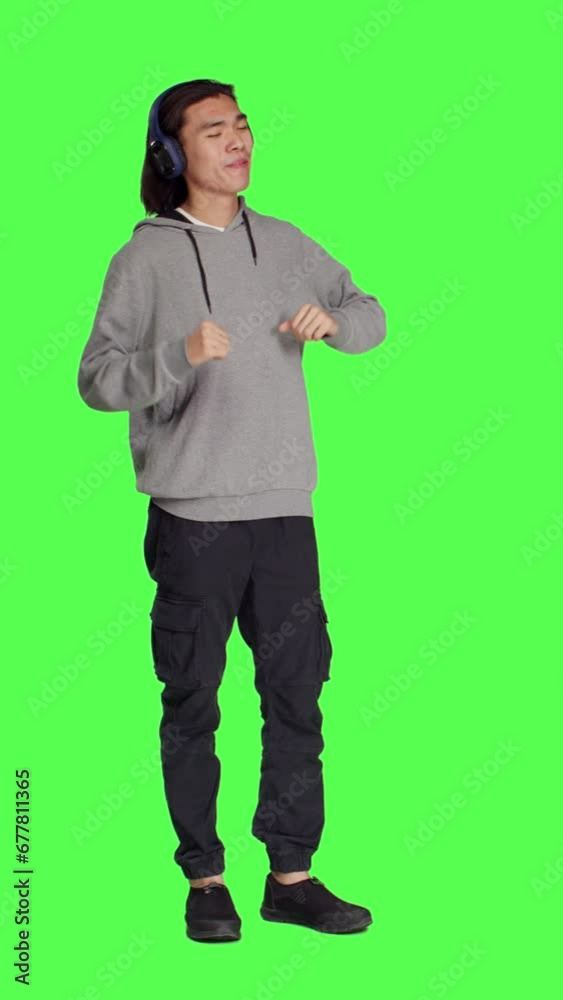 Front view of joyful man does groovy dance moves in studio, feeling funky while he is listening to cool music. Asian male model smiling and enjoying songs on wireless headset, dancing on greenscreen. Front view of joyful man does groovy dance moves in studio, feeling funky while he is listening to cool music. Asian male model smiling and enjoying songs on wireless headset, dancing on greenscreen.