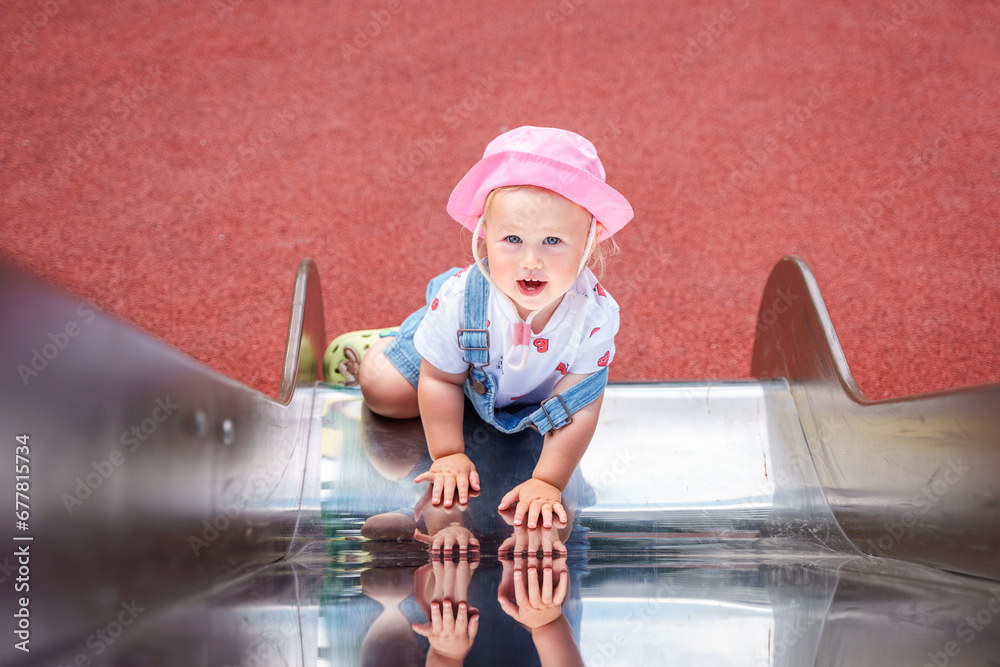 Naklejka premium Cute baby toddler child in denim overalls stands near the slide on the playground,a beautiful concept of caring for children, family and childhood