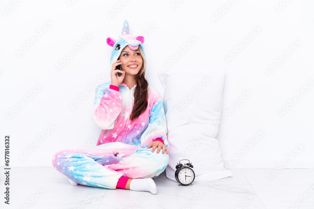 Young Russian girl wearing a unicorn pajama sitting on the floor isolated on white background keeping a conversation with the mobile phone Young Russian girl wearing a unicorn pajama sitting on the floor isolated on white background keeping a conversation with the mobile phone