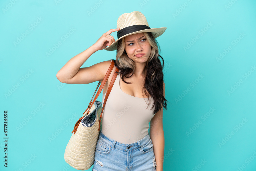 Fototapeta premium Teenager Russian girl with pamel and beach bag isolated on blue background having doubts and with confuse face expression