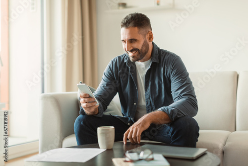 Middle aged man smiles texting via cellphone in living room