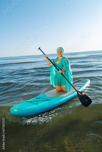 A woman in a turquoise swimsuit with a skirt and a scarf on her head on a SUP board near the sea. © finist_4