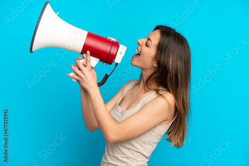 Young caucasian woman isolated on blue background shouting through a megaphone to announce something in lateral position