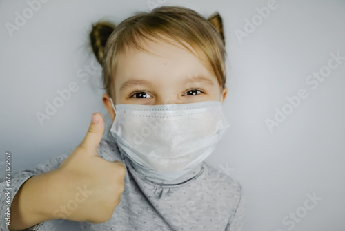 Little girl in a medical mask on a gray background smiles and shows thumb up. Child happy.