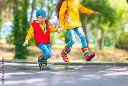 Happy kids girl and boy with umbrella and colorful rubber rain boots playing outdoor and jumping in rainy puddle