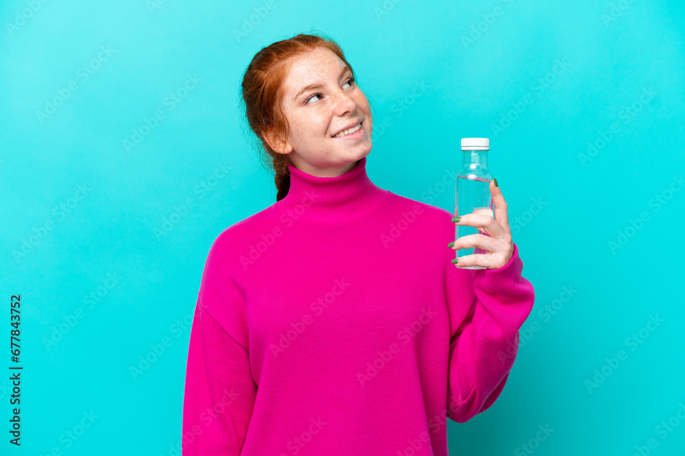 Young caucasian reddish woman with a bottle of water isolated on blue background looking up while smiling Young caucasian reddish woman with a bottle of water isolated on blue background looking up while smiling