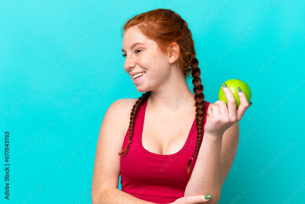 Young reddish woman isolated on blue background with an apple and happy Young reddish woman isolated on blue background with an apple and happy