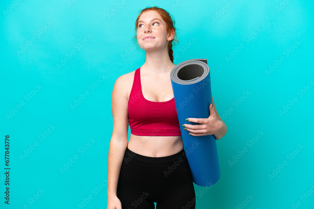 Young sport reddish woman going to yoga classes while holding a mat isolated on blue background and looking up Young sport reddish woman going to yoga classes while holding a mat isolated on blue background and looking up