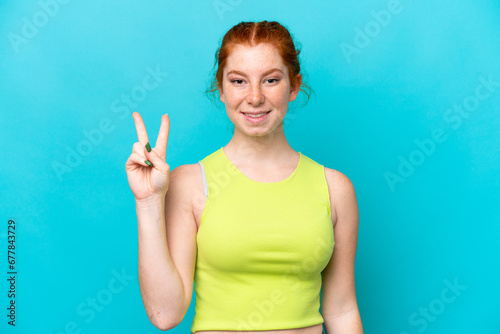 Young reddish woman isolated on blue background smiling and showing victory sign