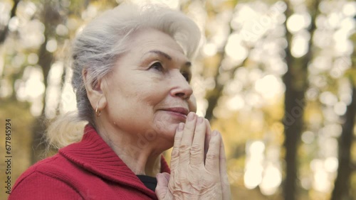 Mature woman with gray hair praying God and starting to amle feeling lightness and purity, charging with positive energy during connection with God outdoor