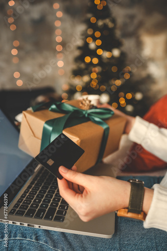 Young woman holding a credit card and a gift box against the background of Christmas decor and gifts, close-up. Christmas and New Year shopping on the Internet, payment by credit card