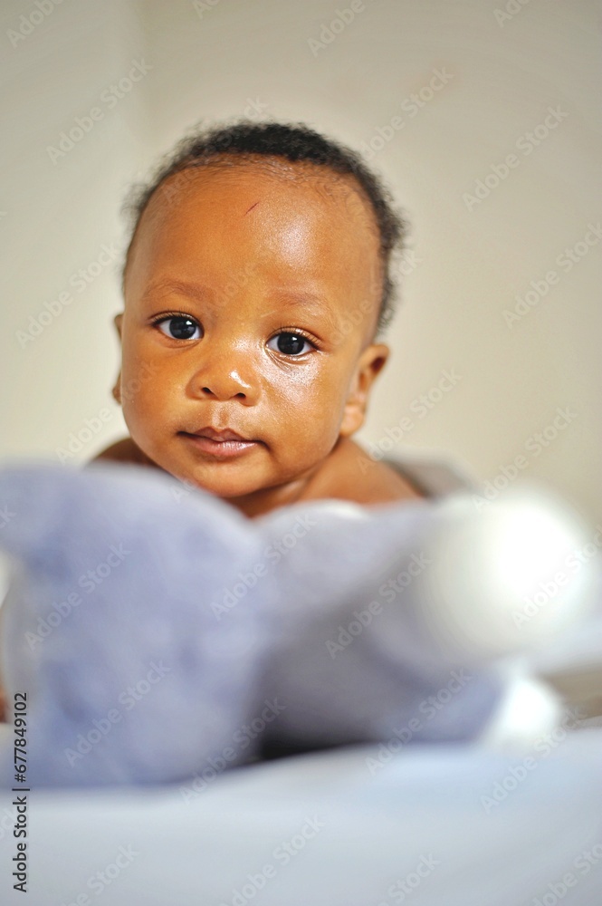 Childcare Concept. Portrait of cute little African  baby wearing bodysuit lying on white bedsheets at home. Black infant child crawling on bed in the bedroom. Selective focus, free copy space