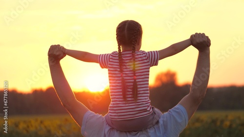 Father holds little daughter on shoulders showing field landscape with sunset