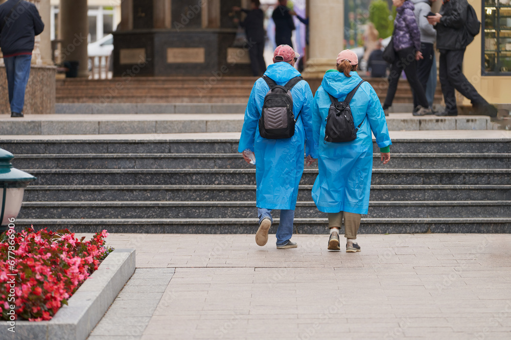 Obraz premium Excursion on a rainy day. A couple in raincoats with backpacks behind their backs on the street of the resort.